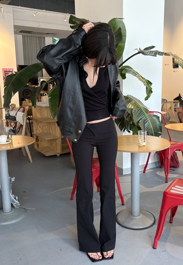 Kvinne iført svart skinnjakke, svart cropped topp og svarte utsvingte bukser som står inne i en kafé med røde stoler og grønne planter.