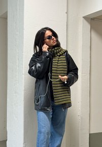 Kvinne med svart lærjakke, stripete grønn skjerf, blå jeans og solbriller, som justerer øretelefonene mens hun står ved en hvit vegg.