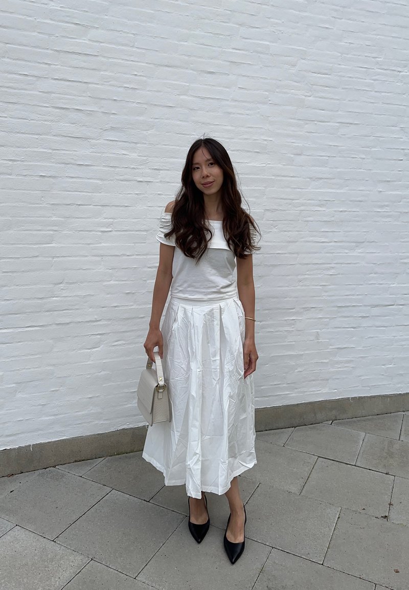 Λευκή εκτός ώμου μπλούζα, πλισέ λευκή φούστα, μαύρες μυτερές γόβες, κρατώντας μια μικρή μπεζ τσάντα, στέκεται πάνω σε γκρίζες πέτρινες πλακίτσες απέναντι από έναν λευκό τοίχο.