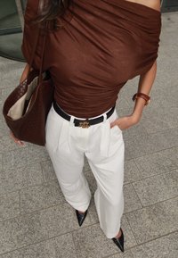 Rudas palaidinė, dėvima ant pečių, baltos klostuotos kelnės su juodu diržu ir aukso spalvos detalėmis, juodi bateliai su aštriais noseliais, ruda pintinė rankinė.