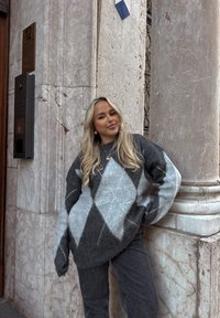 Jeune femme aux longs cheveux blonds appuyée contre un pilier en marbre, portant un pull à motifs losanges gris et noir et un jean foncé.