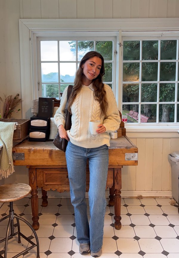 Femme en pull crème et jean tenant une tasse de café, debout à l'intérieur près d'une table en bois et de grandes fenêtres montrant des arbres verts à l'extérieur.