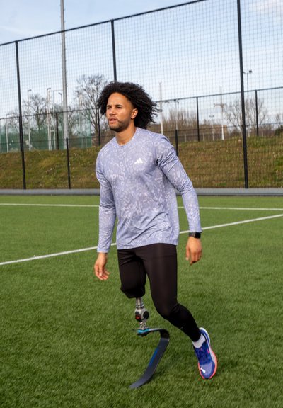 Atleta con una camiseta de manga larga azul claro estampada y leggings negros, utilizando una prótesis de cuchilla para correr, en un campo de césped verde.