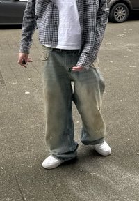 Φαρδιά, ξεθωριασμένα μπλε τζιν με ορατή φθορά και λεκέδες, συνδυασμένα με ένα λευκό μπλουζάκι και ένα καρό πουκάμισο. Λευκά αθλητικά παπούτσια ολοκληρώνουν το σύνολο.