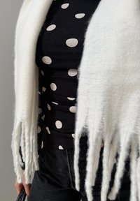 Haut noir à pois blancs sous une écharpe blanche à franges, associé à un pantalon foncé, une main tenant un sac à main noir au niveau de la hanche.