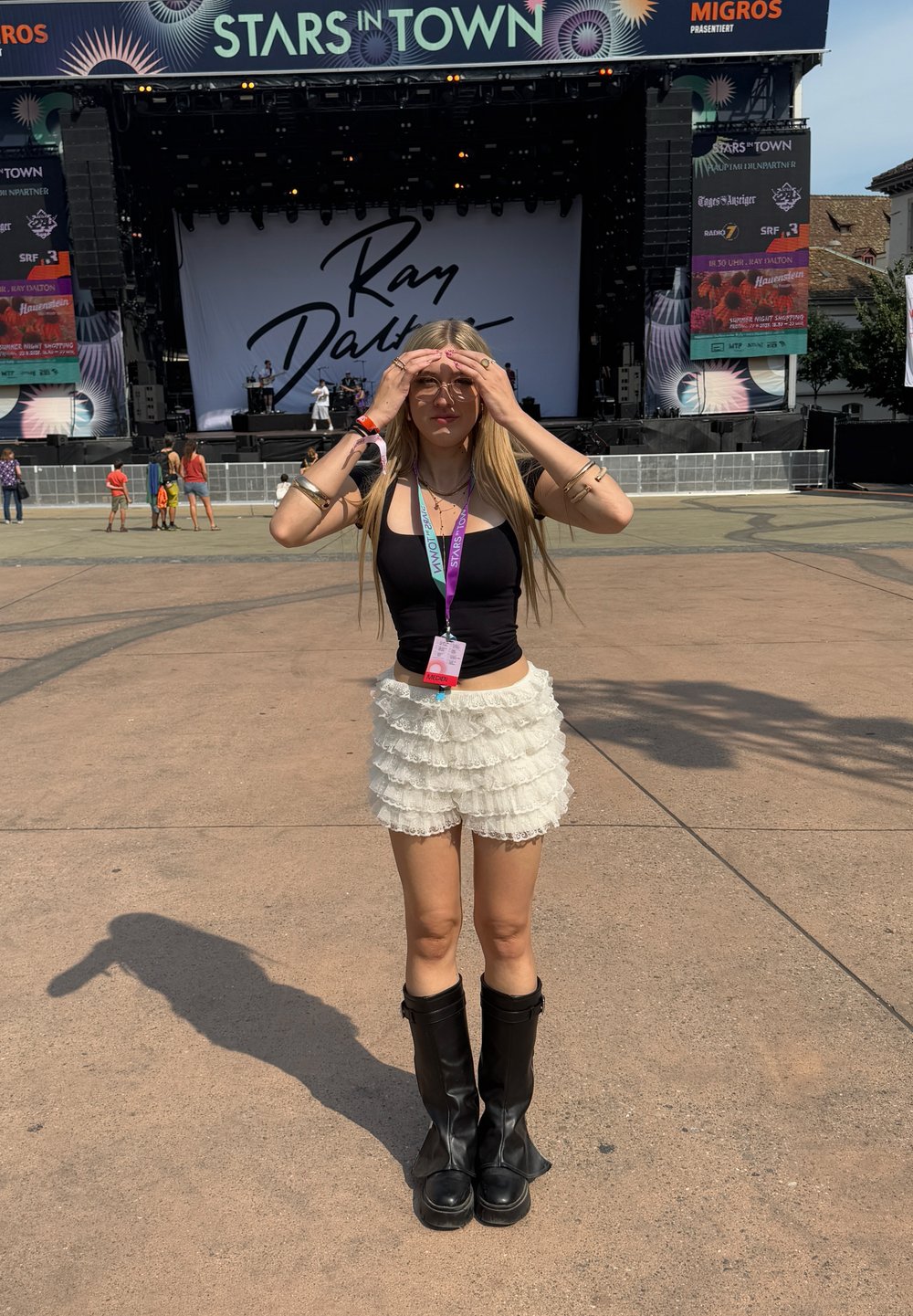 Černý přiléhavý top, bílá vrstvená sukně s volánky a černé kozačky po kolena. Blond vlasy a brýle, stojí před koncertní scénou.