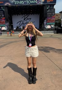 Černý přiléhavý top, bílá vrstvená sukně s volánky a černé kozačky po kolena. Blond vlasy a brýle, stojí před koncertní scénou.