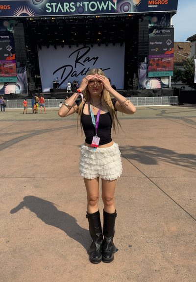 Top negro ajustado, falda blanca con volantes en capas y botas negras hasta la rodilla. Cabello rubio y gafas, de pie frente a un escenario de concierto.