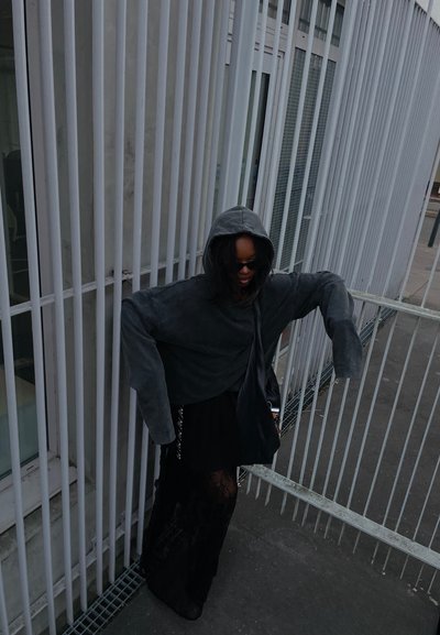 Sudadera gris de gran tamaño con capucha, que presenta mangas largas y un corte relajado, combinada con una falda negra y un bolso cruzado. Fondo texturizado.