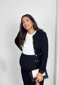 Femme enceinte souriante en cardigan noir et jupe fendue sur le c ôté, chemise blanche, tenant une pochette noir et blanc sur un fond uni.