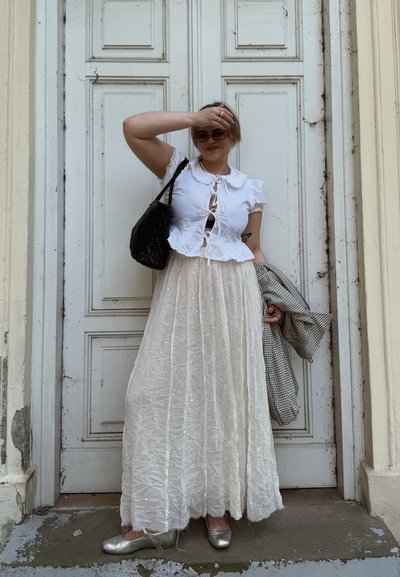 Blusa blanca de manga corta con cuello con volantes y detalle de lazo, combinada con una falda larga y fluida de textura crema y bailarinas plateadas. Bolso negro trenzado.