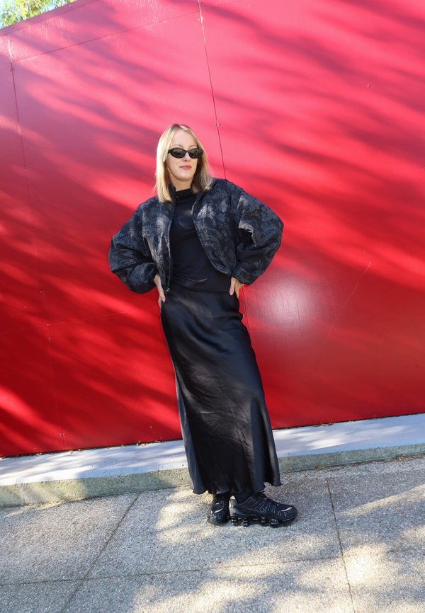 Frau mit schwarzen Sonnenbrillen, einem gemusterten schwarzen Jacket, einem langen schwarzen Rock und schwarzen Schuhen, die draußen an einer roten Wand steht.