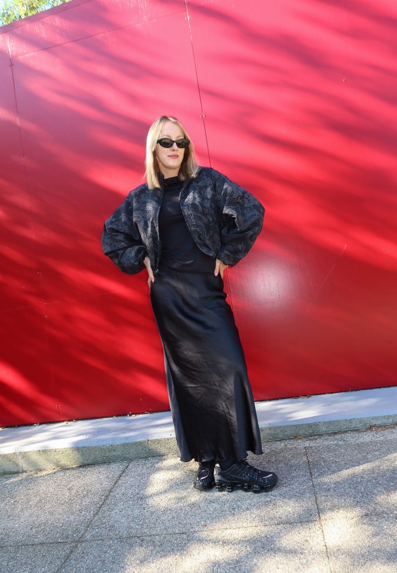 Femme portant des lunettes de soleil noires, une veste noire à motif, une longue jupe noire et des chaussures noires, se tenant devant un mur rouge à l'extérieur.