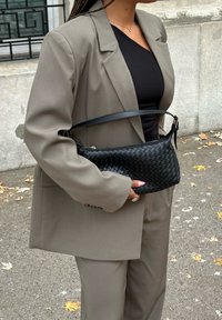 Bolso de cuero negro trenzado con cierre de cremallera y correa para el hombro, sostenido contra un blazer gris de gran tamaño y una blusa negra.