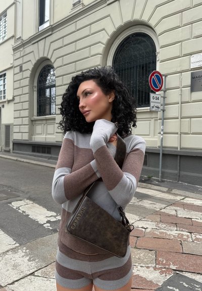 Suéter a rayas en gris y marrón, con mangas largas. Bolsa de hombro marrón con "EXPATRIE" grabado. Entorno urbano con calle adoquinada.