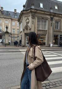 Beige trench coat worn over a grey top, paired with high-waisted blue jeans. A brown tote bag with a textured surface is slung over the shoulder.