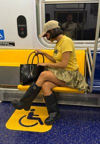 Sorte knelange gummistøvler, camo-skjørt og gul T-skjorte. Håndveske med sølvkjede. Gult T-banested og blåprikket gulv.