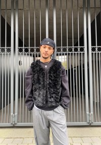Black faux fur vest over a grey sweatshirt, paired with light grey pleated trousers. The background features black metal gates.