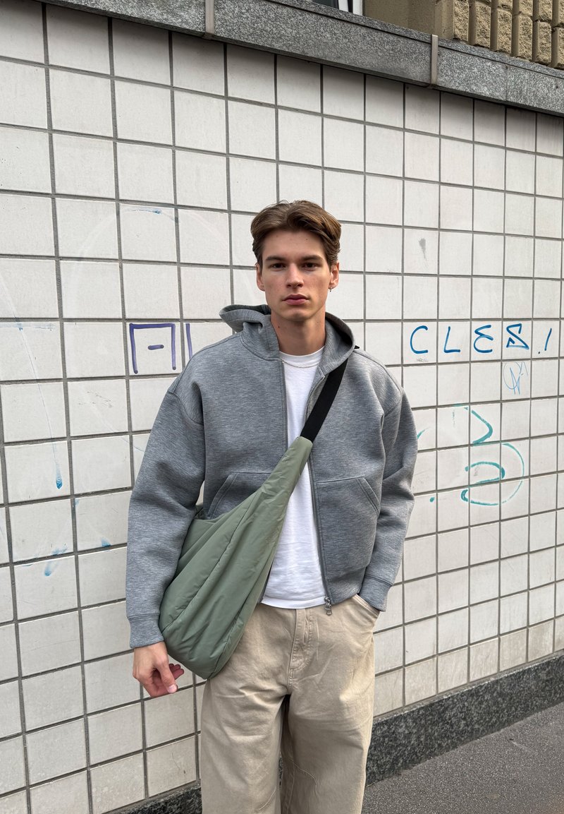 Un sweat à capuche zippé gris sur un t-shirt blanc, un pantalon couleur crème et un sac bandoulière vert. Tissu lisse, coupe décontractée et design subtil.