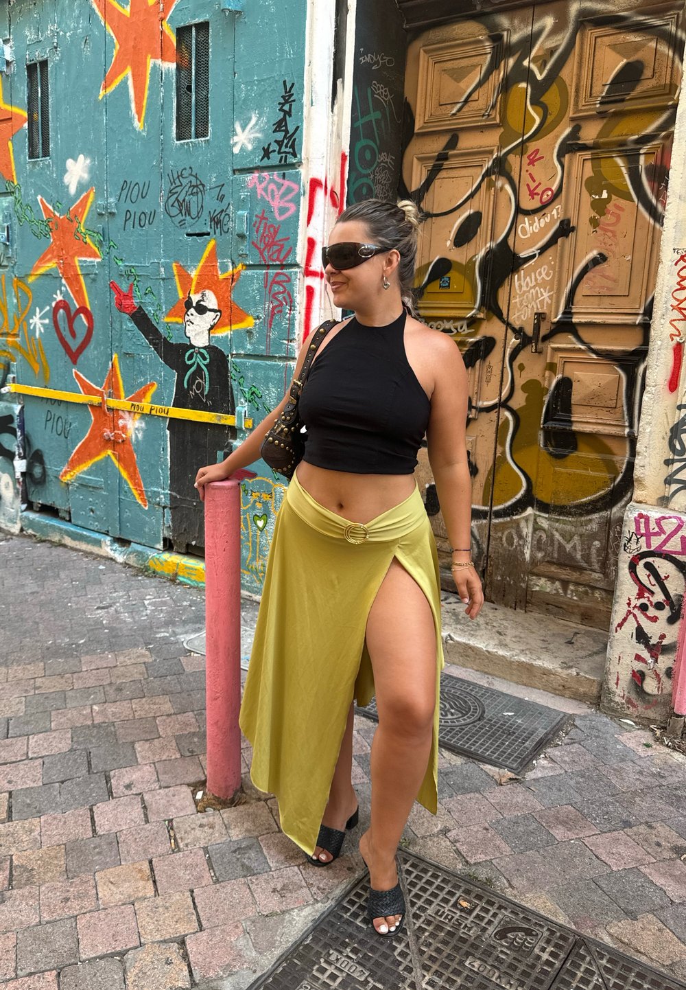 Zwarte fitted crop top, gele wikkelrok met een hoge split en zwarte open teen hakken; staande naast een kleurrijke, met graffiti bedekte muur.