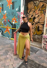 Zwarte fitted crop top, gele wikkelrok met een hoge split en zwarte open teen hakken; staande naast een kleurrijke, met graffiti bedekte muur.
