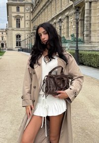 Donna dai lunghi capelli scuri indossa un trench beige sopra un outfit bianco, tiene una borsa marrone ed è in piedi su un sentiero di ghiaia accanto a edifici storici in pietra.