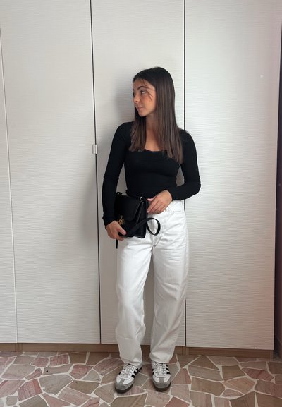 Top negro de manga larga, pantalones blancos de pierna ancha, zapatillas gris y blancas, sosteniendo un bolso negro con un acento dorado. Fondo sencillo.