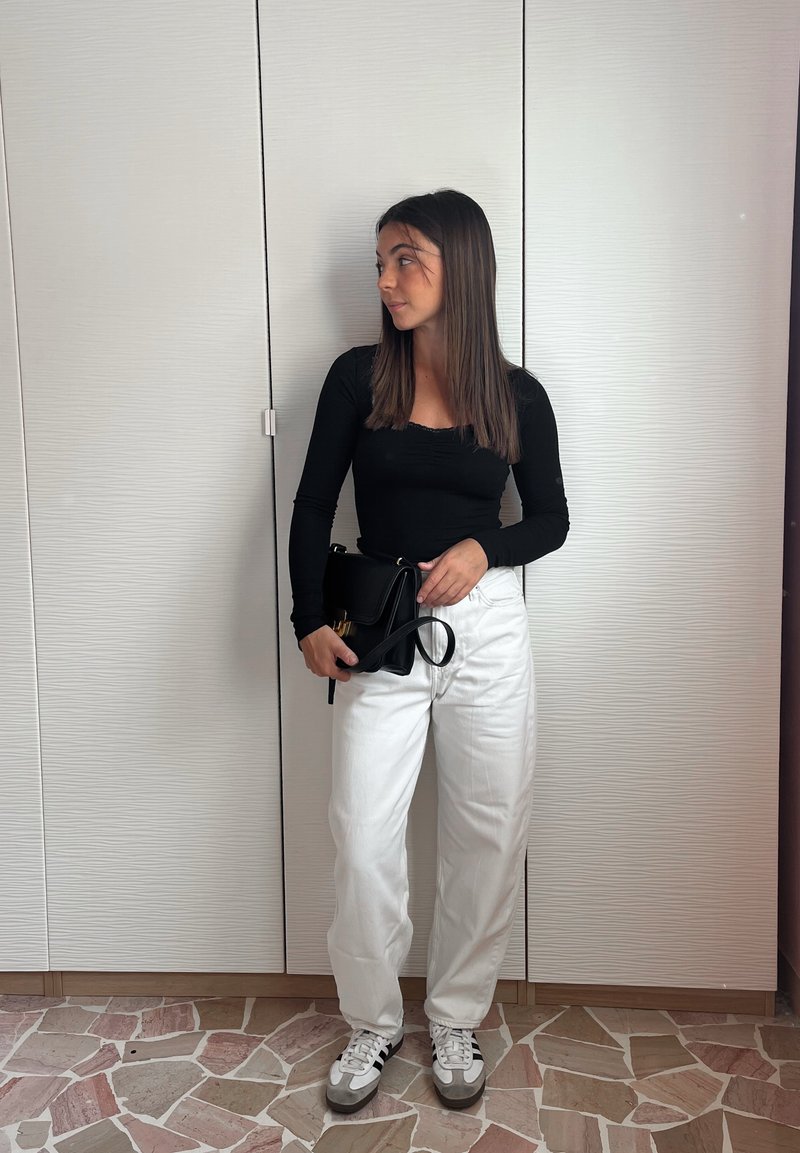 Schwarz langärmliges Oberteil, weiße weite Hose, grau-weiße Sneakers, hält eine schwarze Handtasche mit einem goldenen Akzent. Einfarbiger Hintergrund.