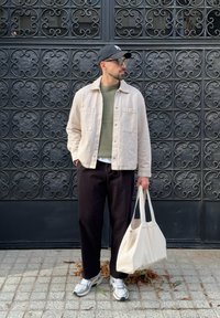 Man in a beige jacket, green sweater, black trousers, and cap holding a white tote bag while standing against an ornate black metal gate.