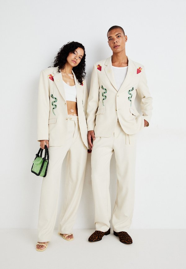 Fatos sob medida em creme com bordados de cobras e corações, combinados com blusas brancas, calças largas e calçado em contraste. Modelo segura uma mala verde.