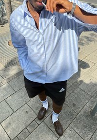 Prugasta modra in bela srajca s kratkimi rokavi, črne hlače s kratkimi nogavicami z belim logotipom Adidas, rjavi loaferji in bele nogavice z modrimi črtami.