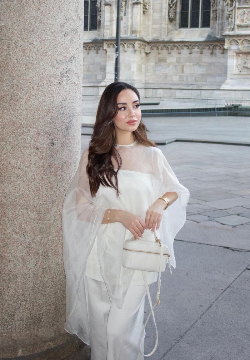 Femme aux longs cheveux foncés portant un top blanc transparent et un pantalon, debout près d'une colonne en pierre, tenant un petit sac à main blanc, bâtiment historique en arrière-plan.