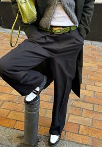 Black wide-leg trousers, green braided belt, grey sweater, green handbag, and black shoes with white accents, posed on a metal post.