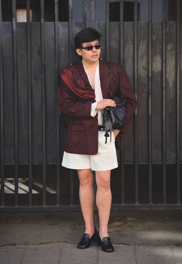Personne portant un blazer bordeaux oversize, un short blanc, des chaussures noires, des lunettes de soleil foncées, tenant un sac noir, debout devant une porte métallique noire.