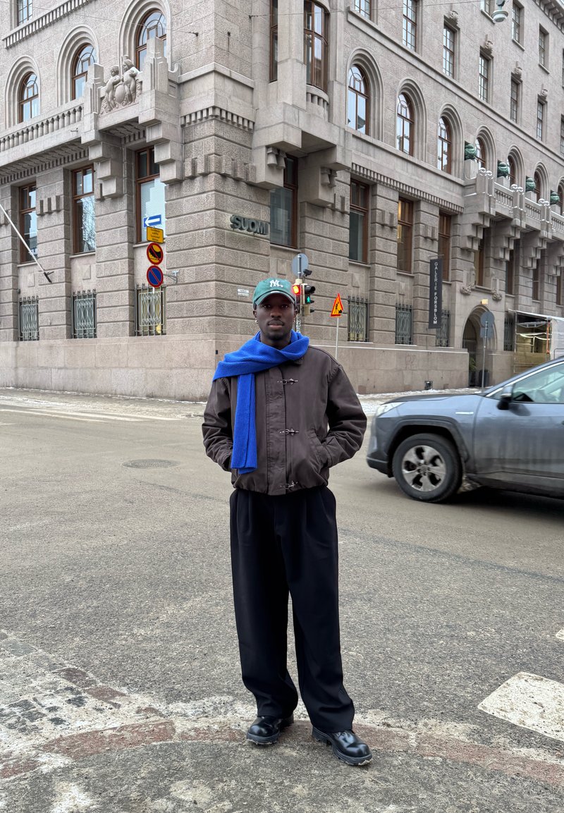 Mann mit schwarzer Jacke, blauem Schal und grüner Mütze steht an einer Straßenecke in der Stadt neben einem steinernen Gebäude mit Bogenfenstern und einem vorbeifahrenden grauen SUV.