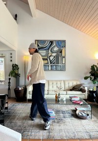 Beige gebreide cardigan, witte shirt, donkere broek en sneakers. De kamer heeft een witte bank, glazen salontafel en moderne kunst. Houten plafond.