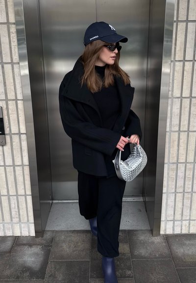 Mujer con abrigo negro, pantalones y gafas de sol, sosteniendo un bolso plateado, saliendo de un ascensor metálico en un entorno urbano.