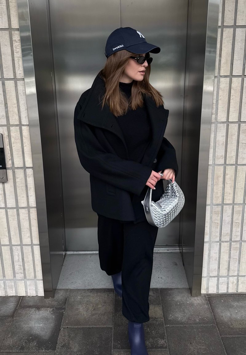 Frau trägt schwarzen Mantel, Hose und Sonnenbrille, hält eine silberne Handtasche und verlässt einen metallischen Aufzug in einer urbanen Umgebung.