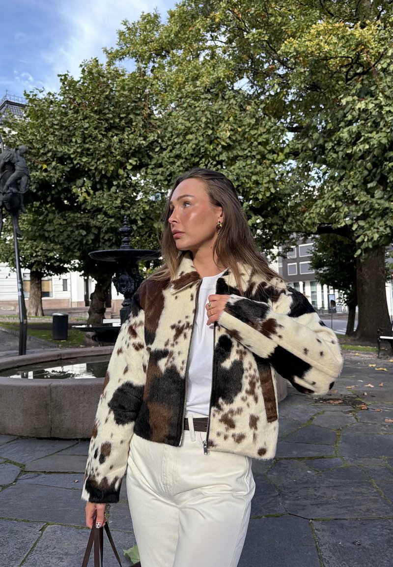 Giacca in pelliccia sintetica con motivo a macchie di mucca in nero e marrone su sfondo crema; presenta una chiusura frontale con zip e una silhouette squadrata.
