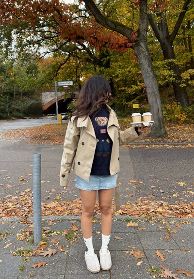 Gabardina beige de doble botonadura sobre un suéter azul marino con un diseño de oso, falda corta de mezclilla, calcetines blancos y zapatos de lona gris claro. Sosteniendo dos tazas de café.