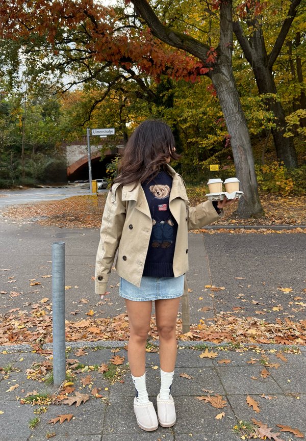 Beige dobbeltbrystet trenchcoat over en marineblå sweater med et bjørnedesign, denim mini skørt, hvide sokker og lysegrå slip-on sko. Holder to kaffekrus.