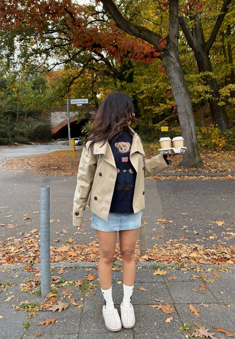 Beige double-breasted trench coat over a navy sweater featuring a bear design, denim mini skirt, white socks, and light grey slip-on shoes. Holding two coffee cups.