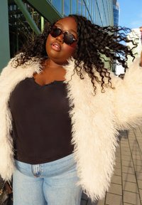 Γυναίκα με σγουρά μαλλιά που φοράει γυαλιά ηλίου, λευκό χνουδωτό παλτό, μαύρο τοπ και τζιν, στέκεται έξω κοντά σε σύγχρονα κτίρια κάτω από τον ήλιο.