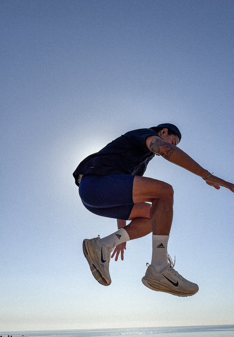 Jeune homme avec des tatouages et des vêtements de sport sautant en l'air sur fond de ciel clair et d'océan.