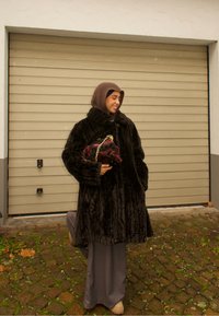 Long manteau en fourrure sombre avec tissu texturé, col montant et gros boutons. Associé à une pochette à motif floral et un pantalon gris ample.