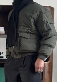 Personne portant une veste matelassée vert olive avec des poches à rabat, une écharpe noire, un pantalon noir et des bagues argentées aux doigts à l'intérieur.