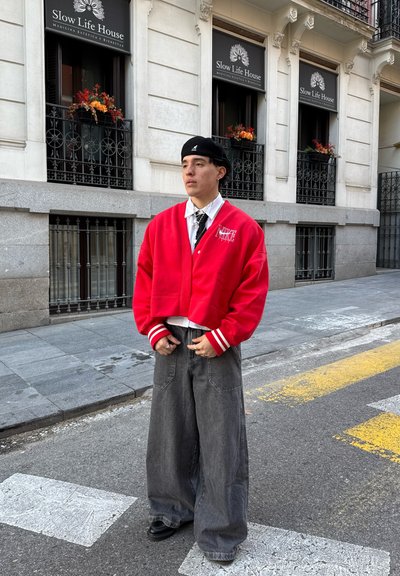 Chaqueta roja corta con rayas blancas, camiseta negra, pantalones anchos grises y zapatos negros. Entorno urbano con arquitectura vintage.