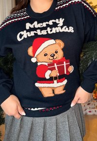 Maglione blu scuro con un orso festivo in un completo da Babbo Natale, che tiene un regalo rosso. Decorato con un bordo decorativo a motivi sul collo e sull'orlo.