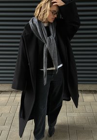 Oversized black coat with a soft grey scarf draped around the neck, paired with black wide-leg jeans and pointed black shoes. Textured surface.