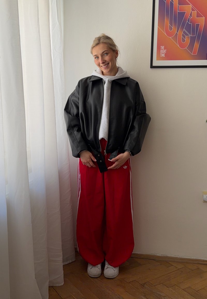 Jeune femme souriante, portant une veste en cuir noire, un sweat à capuche gris, un pantalon large rouge Adidas et des baskets blanches, debout à l'intérieur près d'une fenêtre.